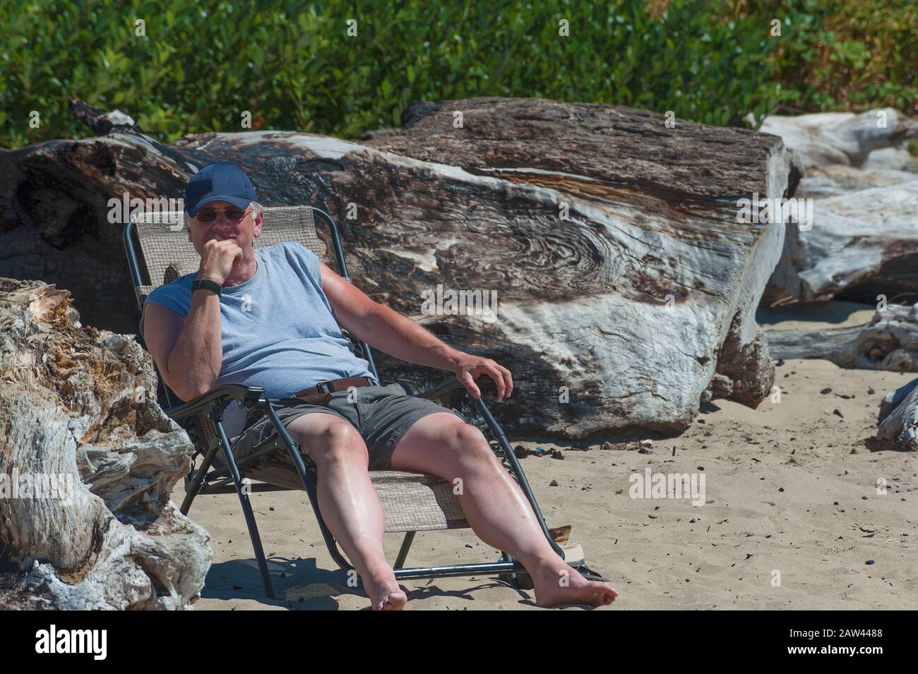 Indossando un tappo a sfera e occhiali da sole, un uomo anziano sale su una chaise pieghevole vicino a grande driftwood sulla spiaggia al Siletz Bay Park a Lincoln City, Oregon Foto Stock