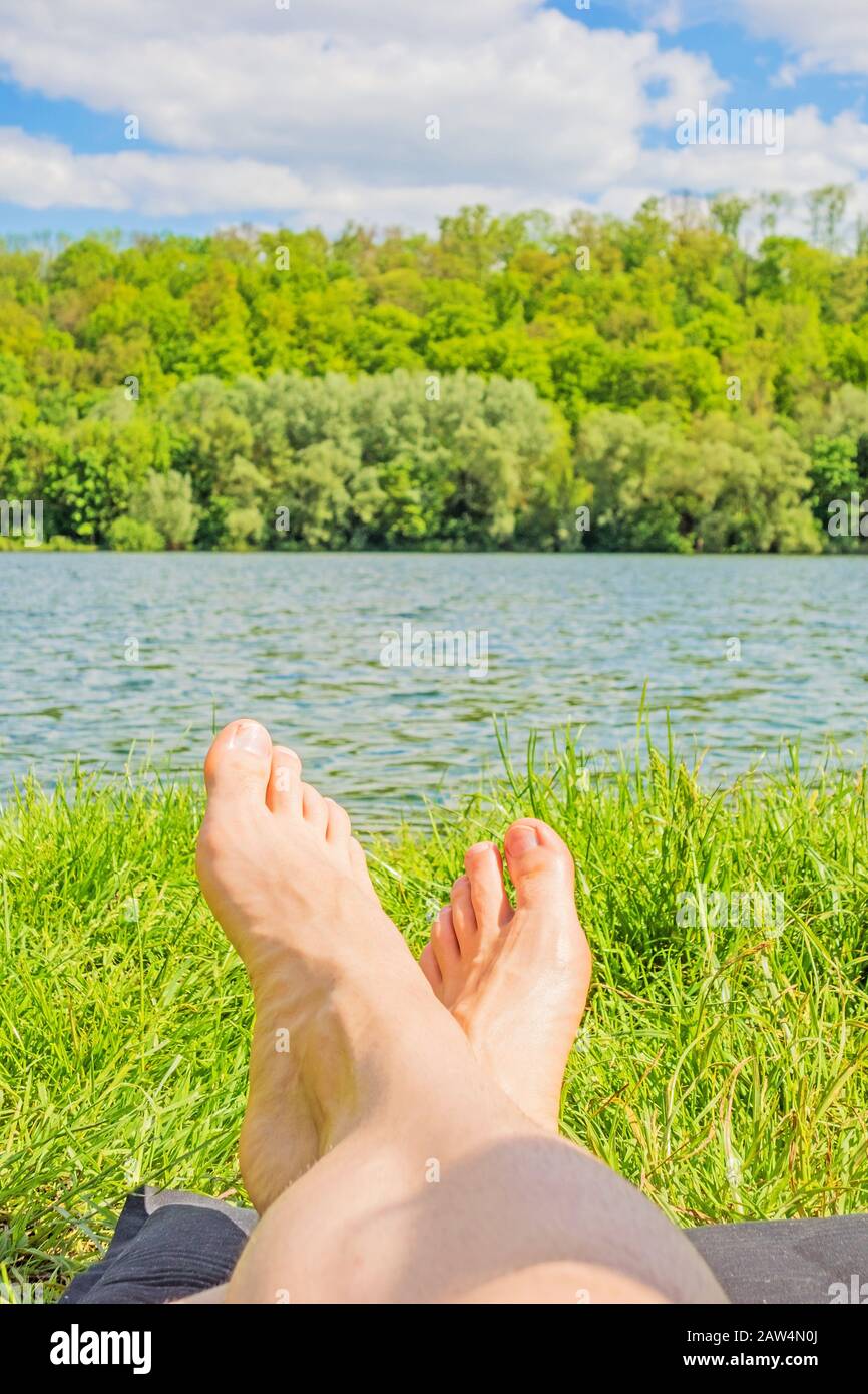 piedi maschili al lago - uomo rilassante al lago sul tappeto campeggio - verde foresta / alberi sullo sfondo Foto Stock