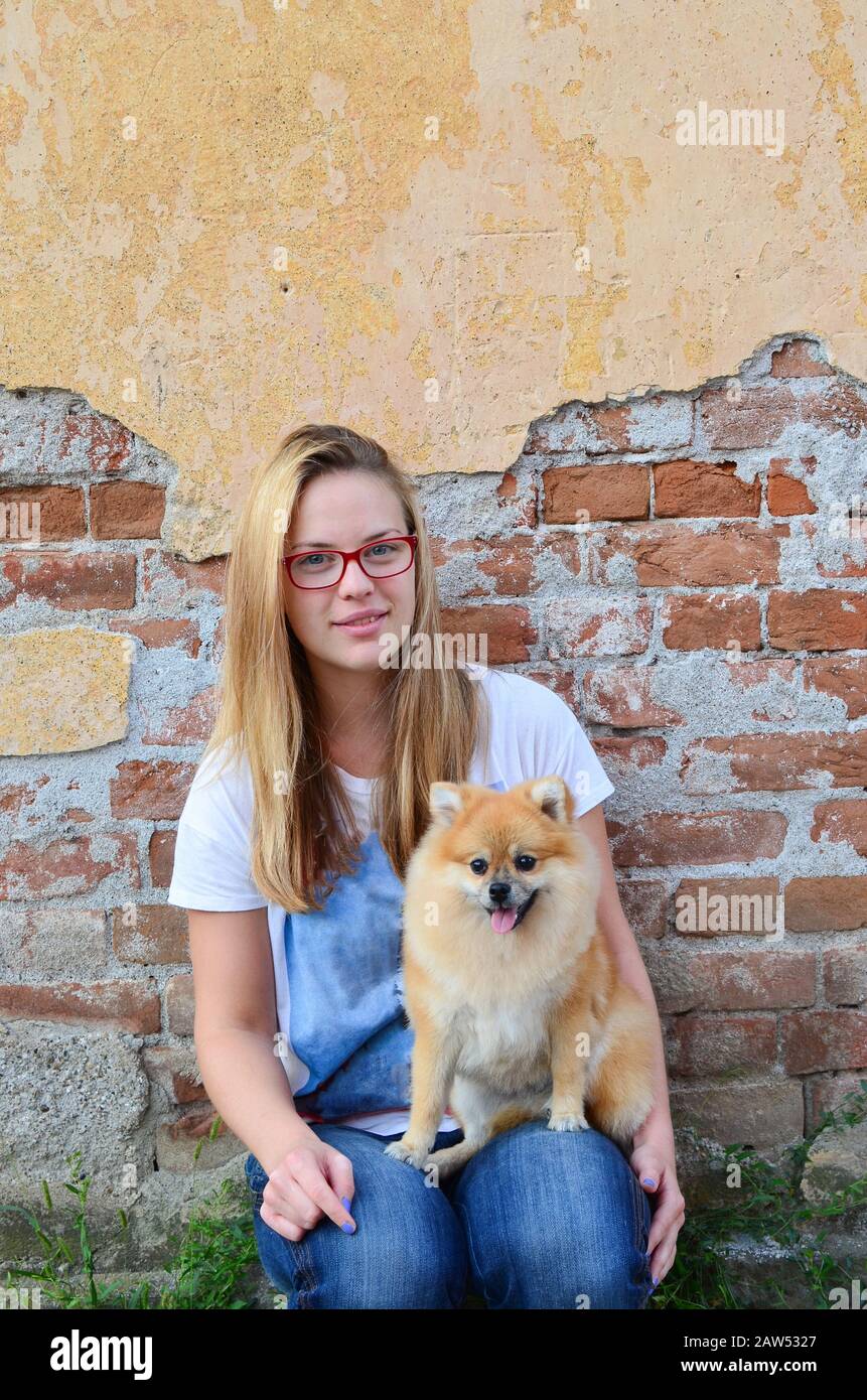 Ragazza giovane, cute accanto porta che indossa occhiali rossi, jeans e t-shirt, posando con il suo animale domestico di fronte a grunge muro di mattoni Foto Stock