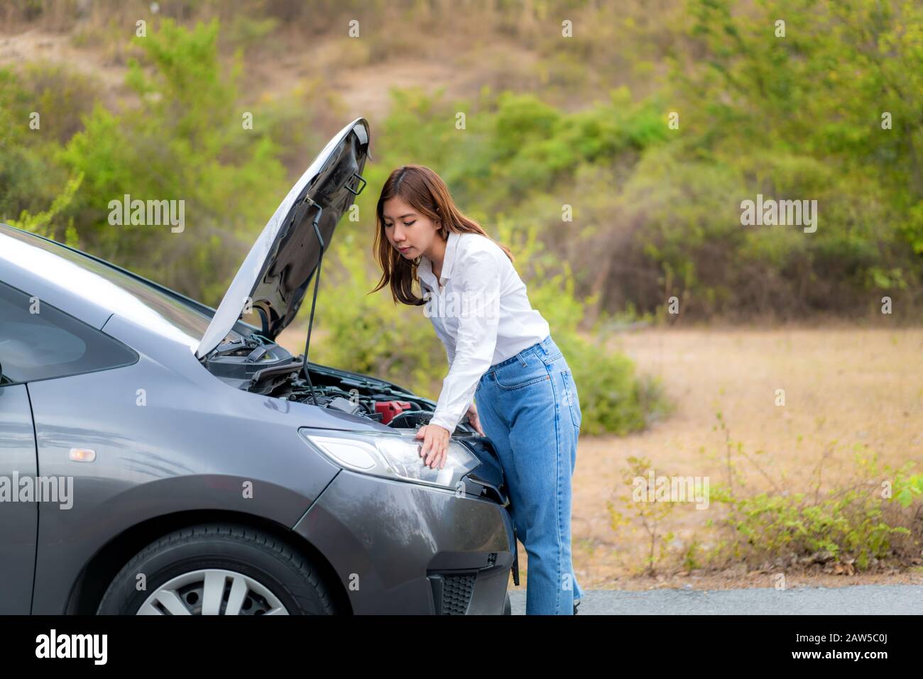 Asian giovane infelice donna che ispeziona motore rotto auto di fronte al cofano aperto rotto auto su strada In Attesa di assistenza stradale servic Foto Stock