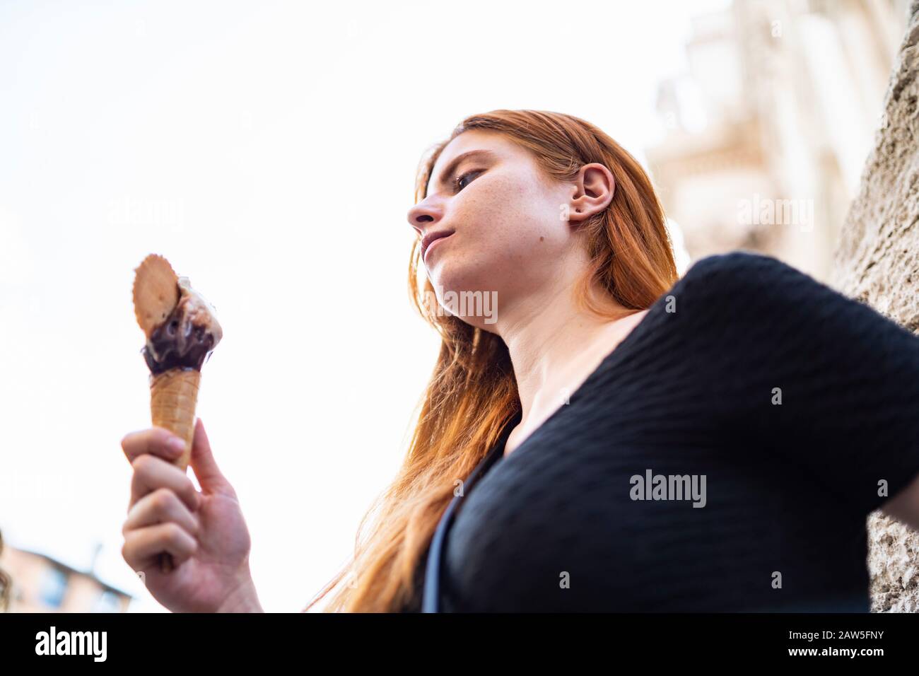 Da sotto vista laterale di giovane femmina casual con capelli rossi che tengono in mano fusione gelato cono con cioccolato e biscotto mentre riposa sul re città Foto Stock