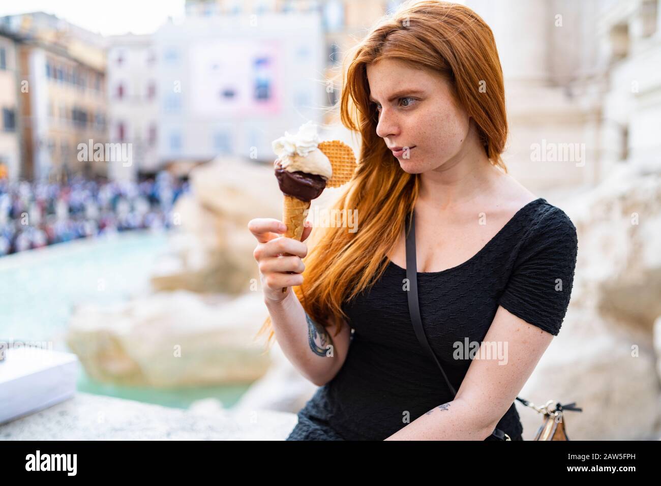 Stupita giovane femmina zenzero in abito casual guardando grande cono gelato mentre si siede sulla strada della città in calda giornata estiva Foto Stock