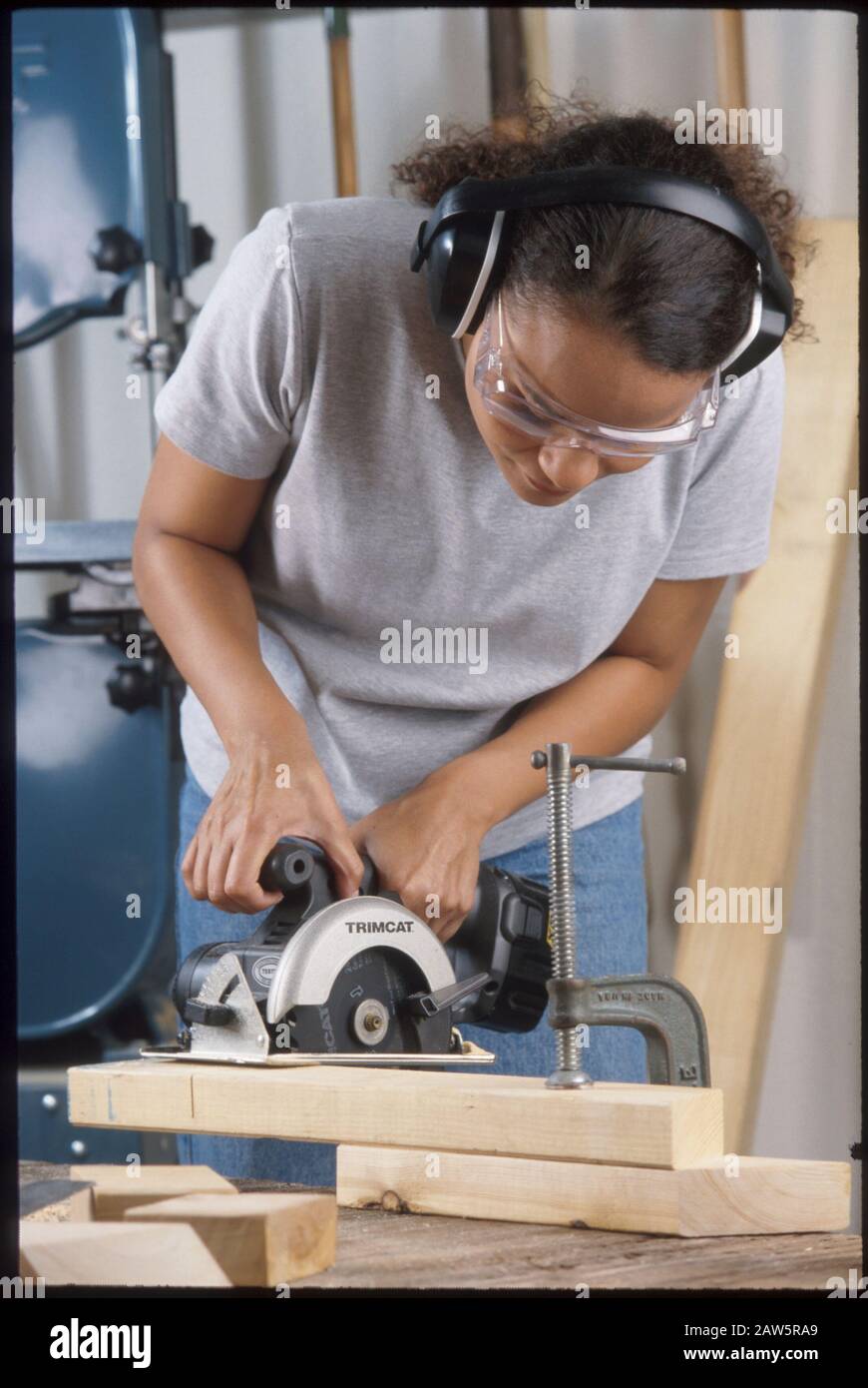 La giovane donna afroamericana indossa occhiali di sicurezza e protezione per l'orecchio mentre taglia con sega circolare senza fili nel suo laboratorio di garage ad Austin, Texas. Foto Stock
