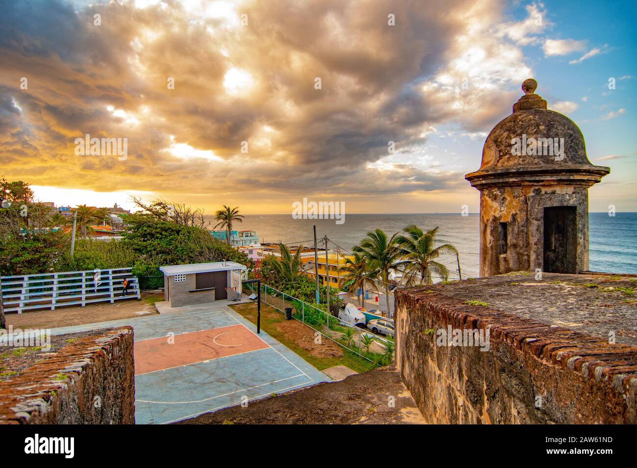 Una torre di osservazione al Castillo San Cristobal Fort si affaccia sulle case colorate di la Perla e un campo da basket con un tramonto colorato. Foto Stock