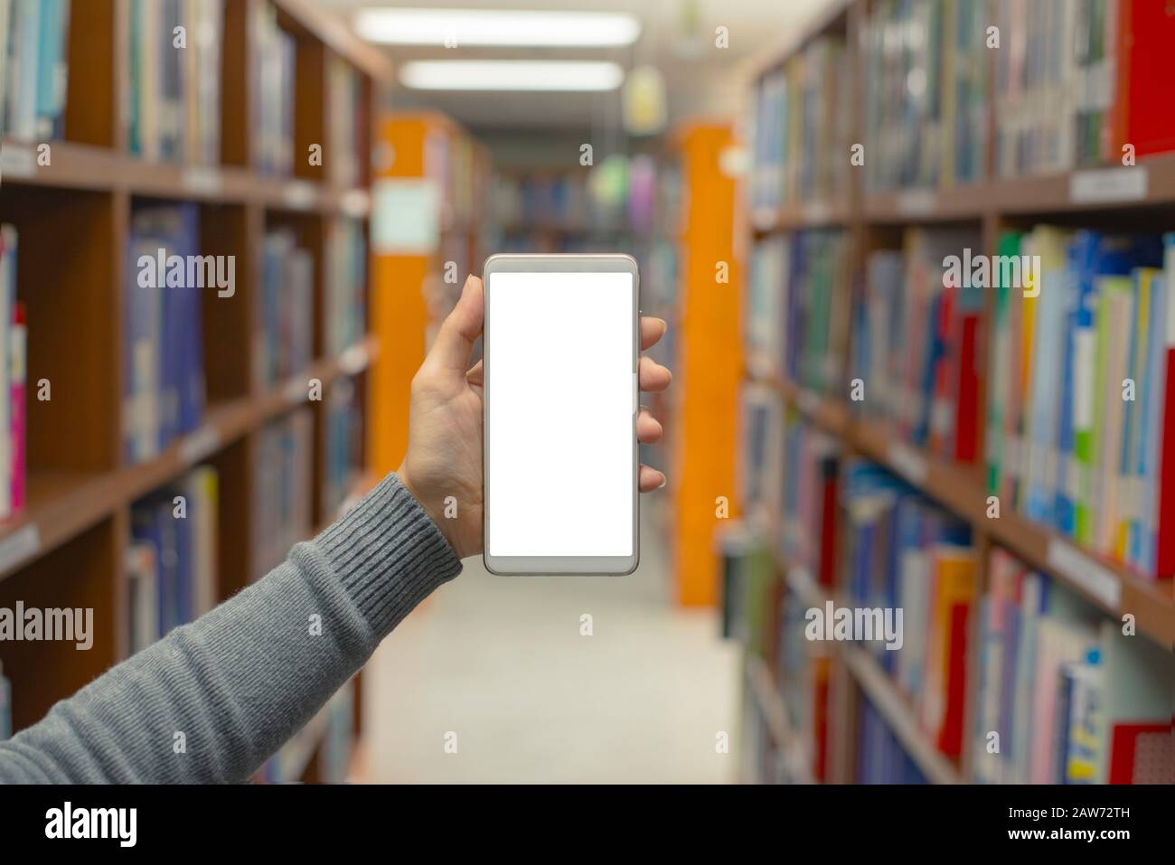 Mani ravvicinate che tengono lo smartphone mobile con sfondo di libri sullo scaffale e stack in biblioteca, concetto di istruzione. Foto Stock