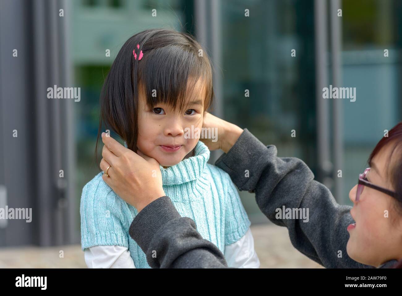Giovane madre asiatica che regola i capelli delle sue figlie intorno ad un maglione dolcevita mentre sorride prettily alla macchina fotografica Foto Stock