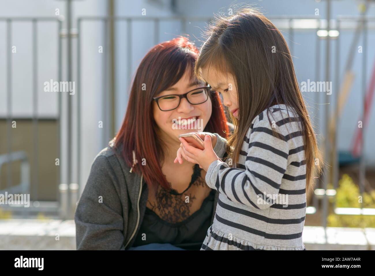 Piccola bambina che gioca con un telefono cellulare tenuto in un abbraccio amorevole da sua giovane madre sorridente all'aperto in città Foto Stock