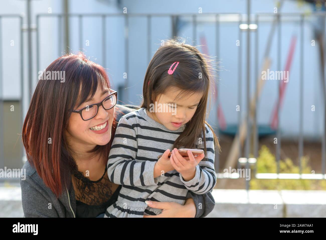 Piccola bambina che gioca con un telefono cellulare tenuto in un abbraccio amorevole da sua giovane madre sorridente all'aperto in città Foto Stock
