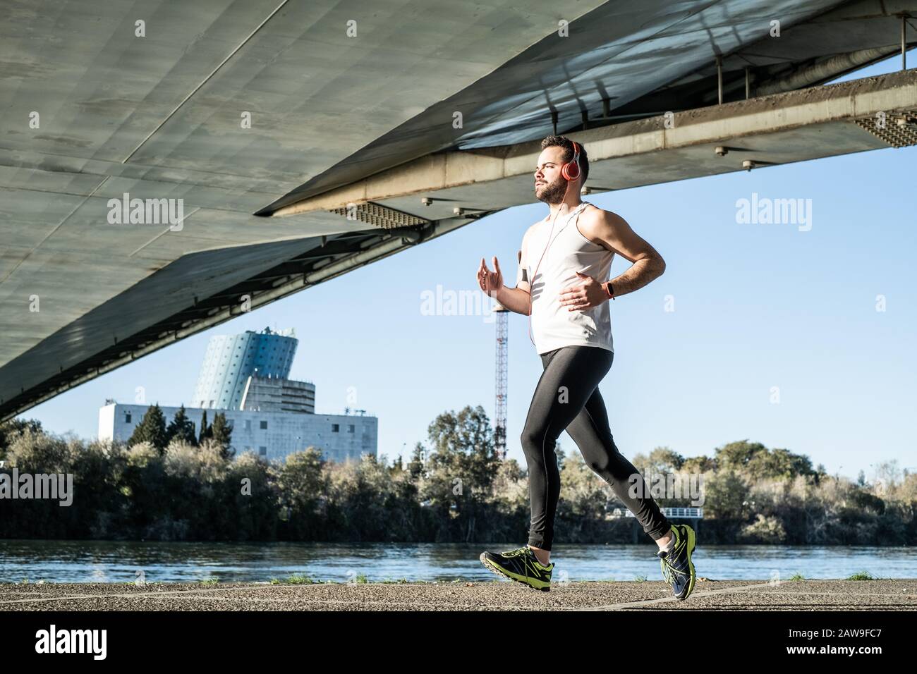 giovane uomo che corre in una camicia bianca attraverso un ponte. Sta ascoltando musica e ha un casco. Foto Stock