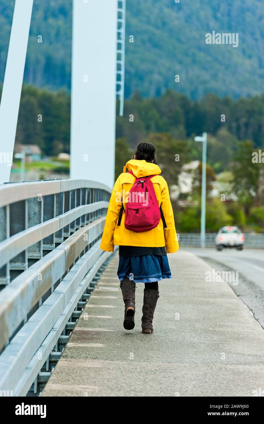 Le donne native dell'Alaska indossano una giacca da pioggia gialla dai colori vivaci e uno zaino arancione che cammina sul ponte o'Connell di Sitka, Alaska, Stati Uniti. Foto Stock