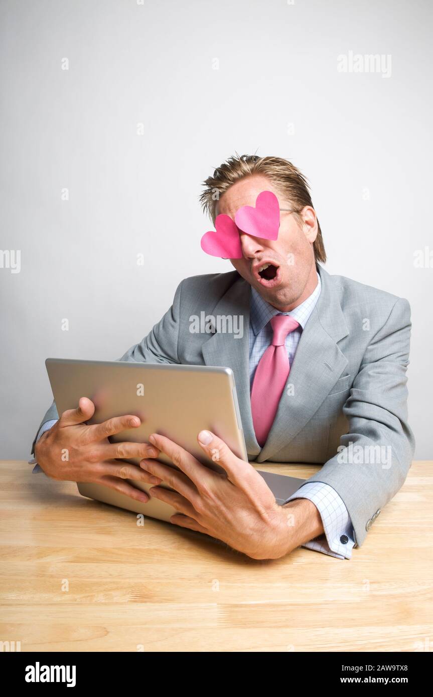 L'operaio dell'ufficio accecato dai cuori rosa che parlano allo schermo mentre abbraccia il calcolatore del laptop sulla sua scrivania Foto Stock