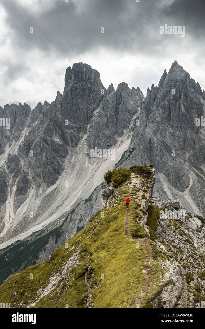 Uomo con giacca rossa in piedi su un grado, dietro di lui cime di montagna e rocce appuntite, nuvole drammatiche, Cimon la Croda liscia e il gruppo Cadini Foto Stock