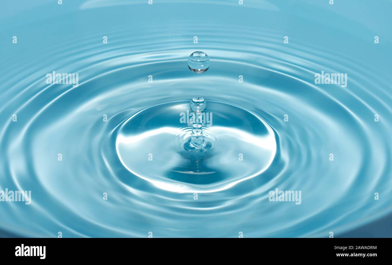 una goccia d'acqua cade con uno spruzzo in acqua chiara e blu che forma cerchi sull'acqua. Foto Stock