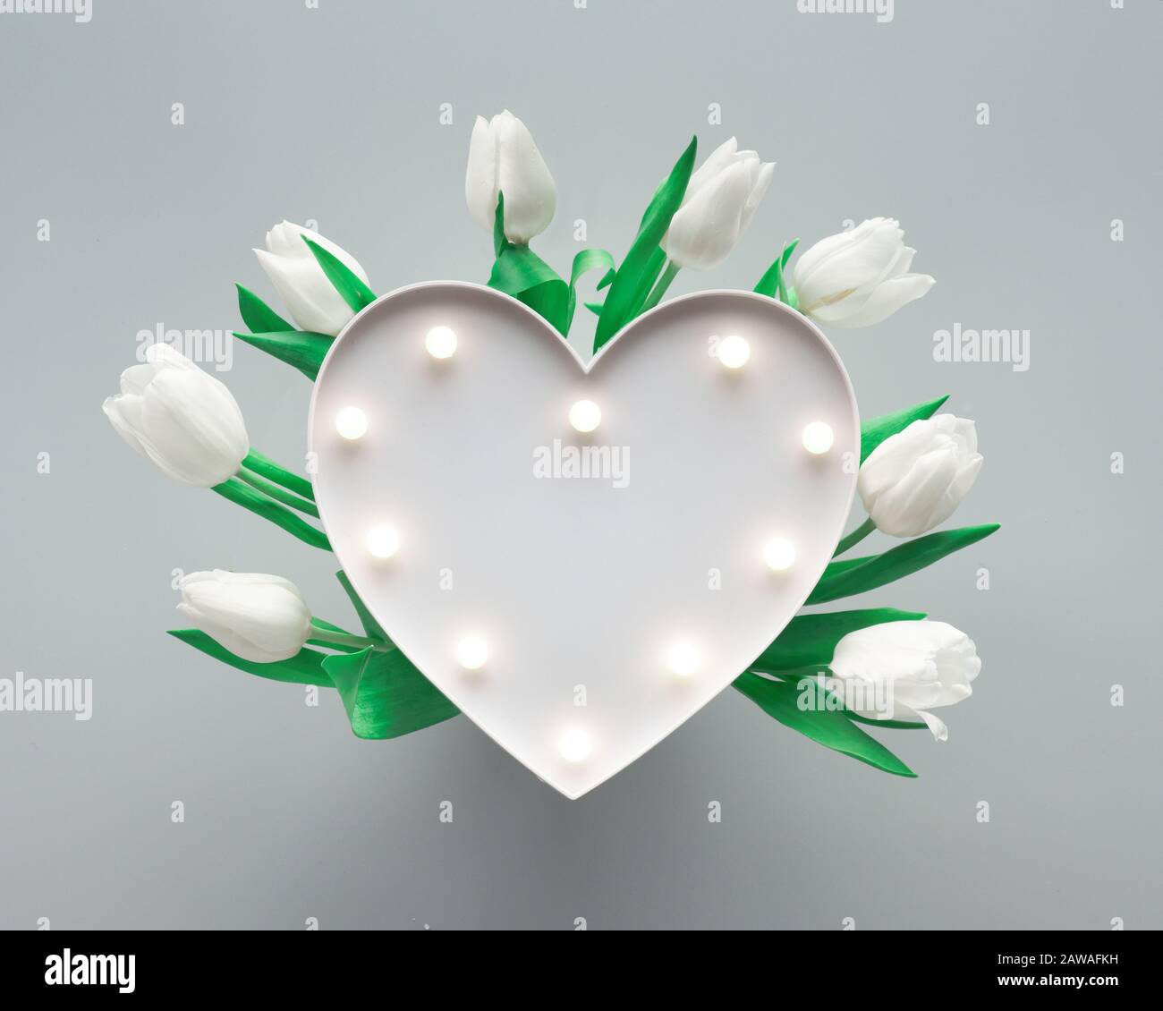 Sfondo a molla con lavagna luminosa a forma di cuore illuminata con luci.  Il cuore è circondato da tulipani bianchi su carta grigio argento. Piatto l  Foto stock - Alamy