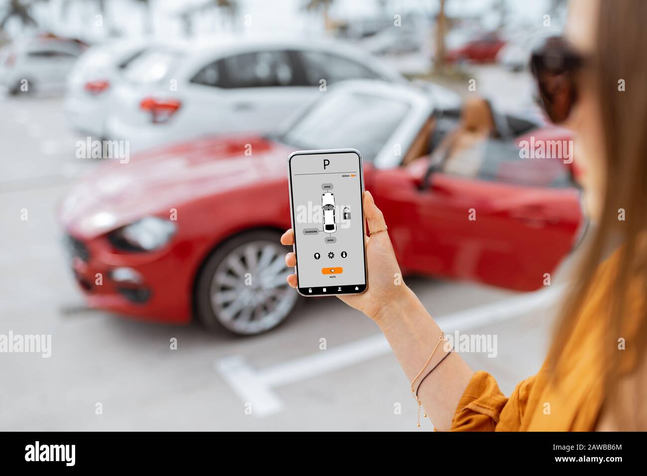 Donna che controlla l'auto con l'applicazione mobile mentre si trova vicino al veicolo al parcheggio, vista ravvicinata sullo smartphone con interfaccia app Foto Stock
