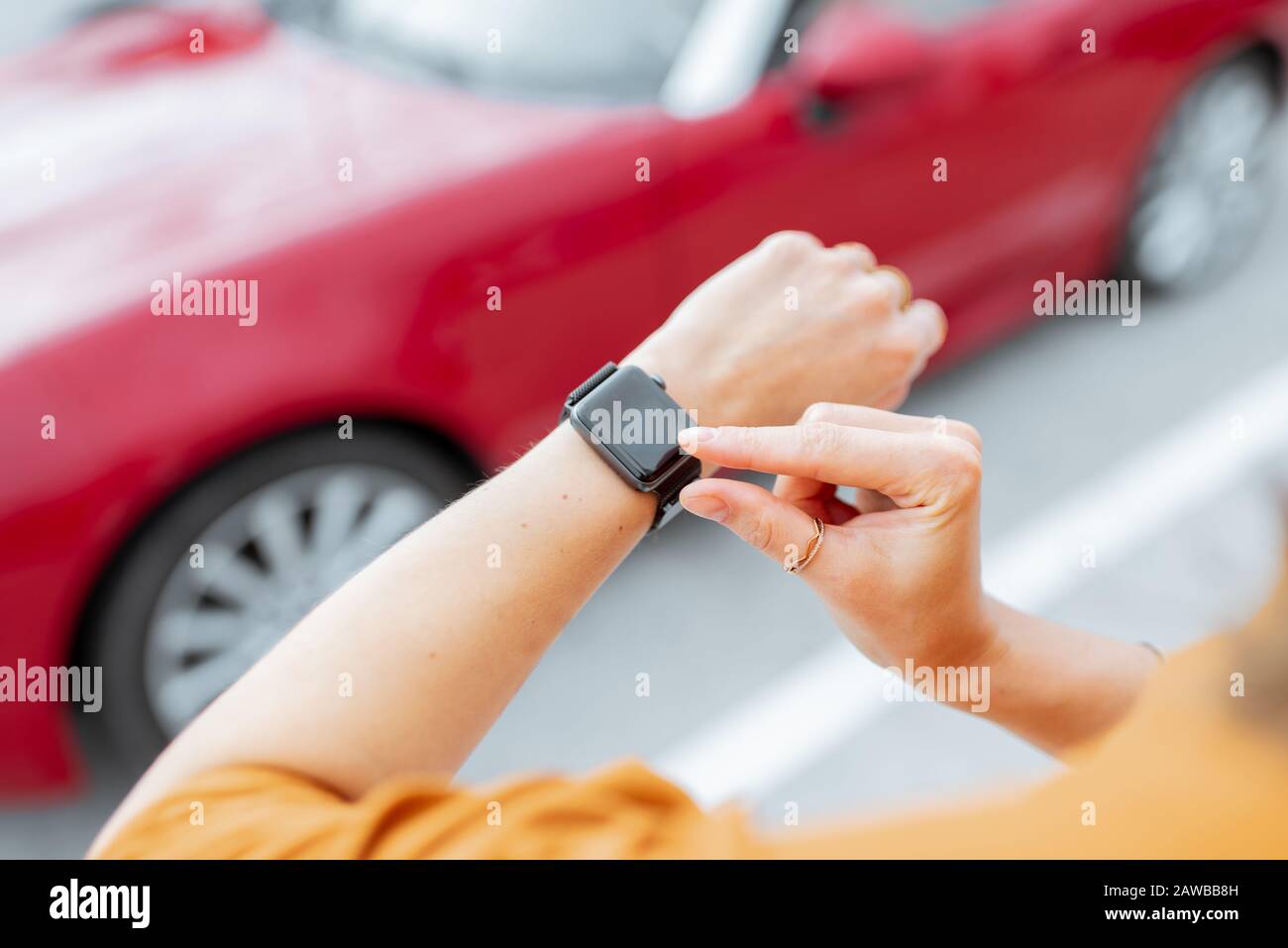 Donna con smartwatch al parcheggio con veicolo rosso sullo sfondo, vista ravvicinata sull'orologio con schermo nero. Controllo dell'auto con un concetto di orologio intelligente Foto Stock