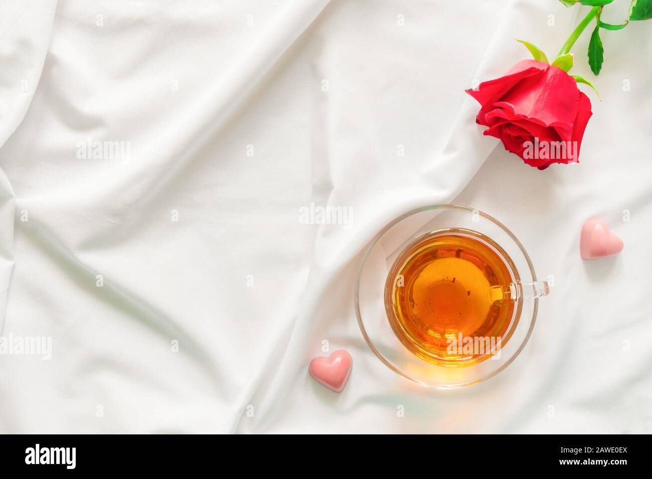 Vista dall'alto della tazza di tè con rosa rossa e caramella a forma di cuore sul letto bianco con spazio copia. Concetto di sorpresa romantica Foto Stock