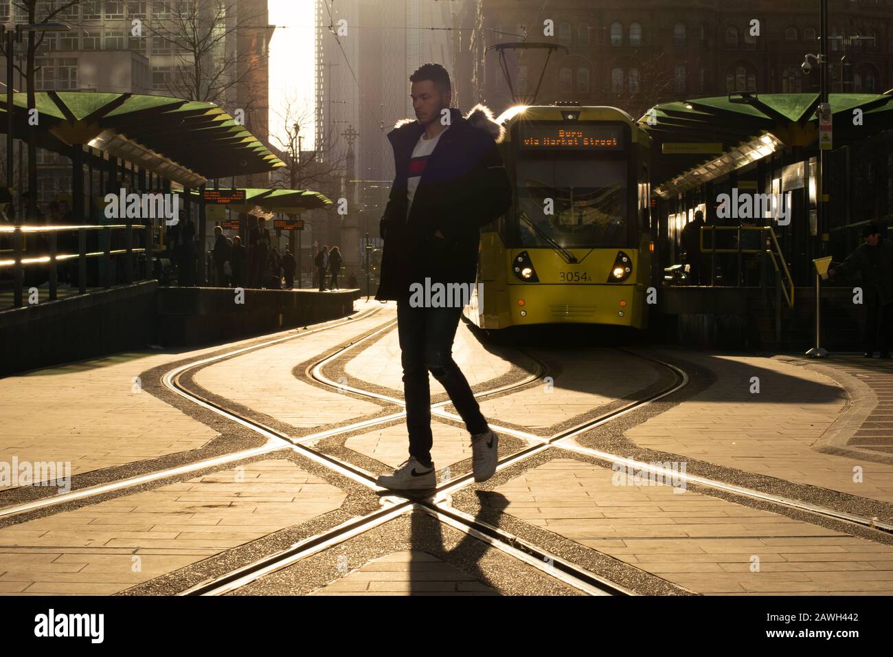 Un uomo cammina attraverso la strada alla fermata del tram Metrolink St Peter's Square Manchester con un tram giallo sullo sfondo. Foto Stock