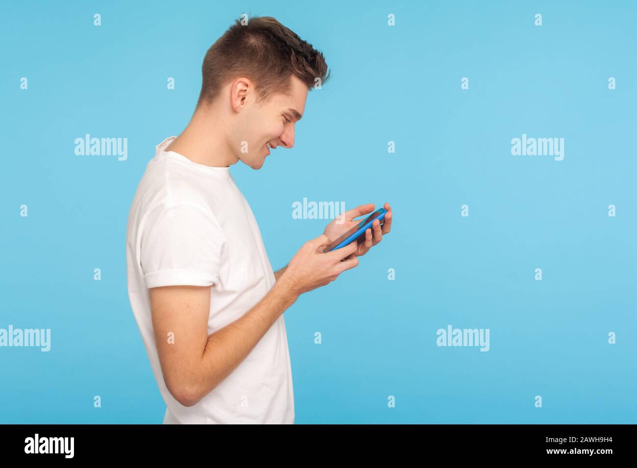 Vista laterale di uomo felice in t-shirt bianca utilizzando cellulare con allegra espressione soddisfatta, ridendo al messaggio in rete sociale, mobile communicatio Foto Stock