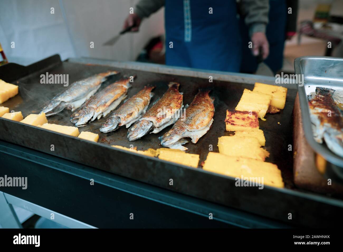 Profondità di campo poco profonda (fuoco selettivo) immagine con un uomo che cucina trote pesce e polenta su una stufa all'aperto in una giornata fredda. Foto Stock