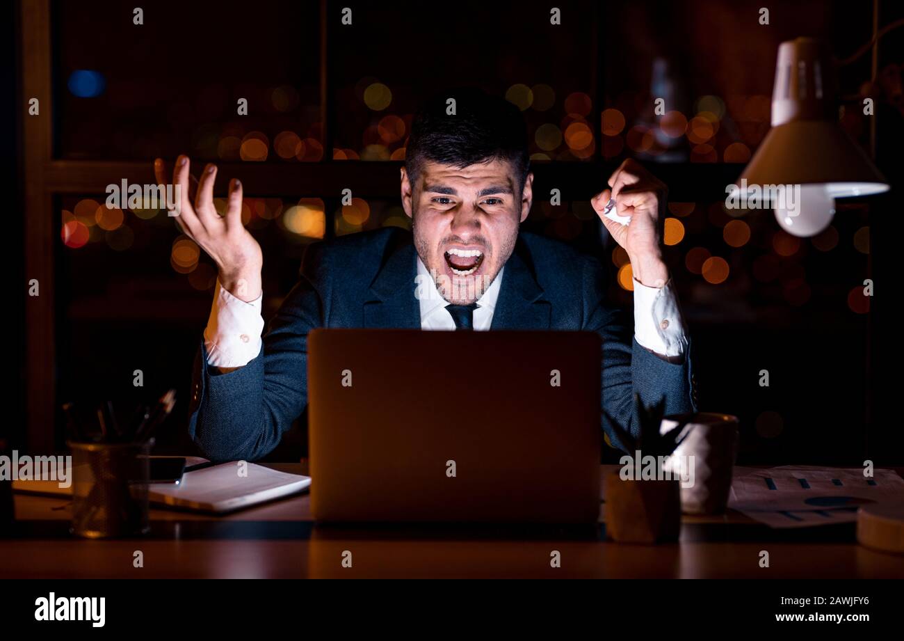 Uomo D'Affari Pazzo Al Laptop Gridando Seduto In Ufficio Di Notte Foto Stock