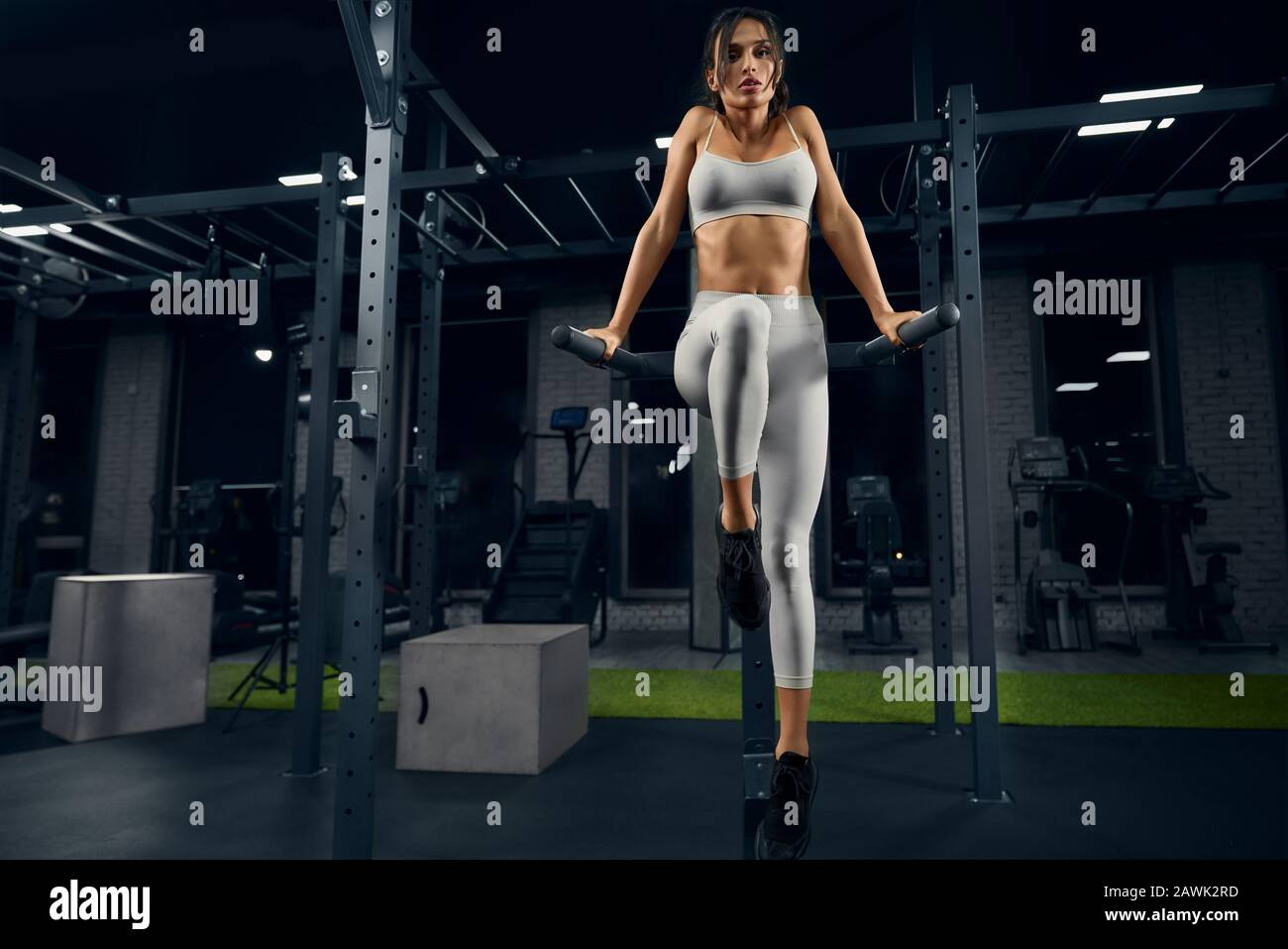 Da sotto vista di atletica giovane donna con il corpo perfetto facendo dips tricipiti sui bar paralleli in palestra. Bella bruna femmina in leggero allenamento sportivo in un'atmosfera buia. Concetto di fitness. Foto Stock