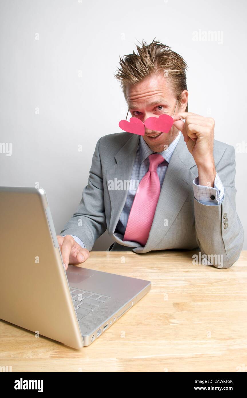 Lavoratore dell'ufficio seduto alla sua scrivania che abbassa gli occhiali rosa del cuore per guardare il suo computer portatile Foto Stock
