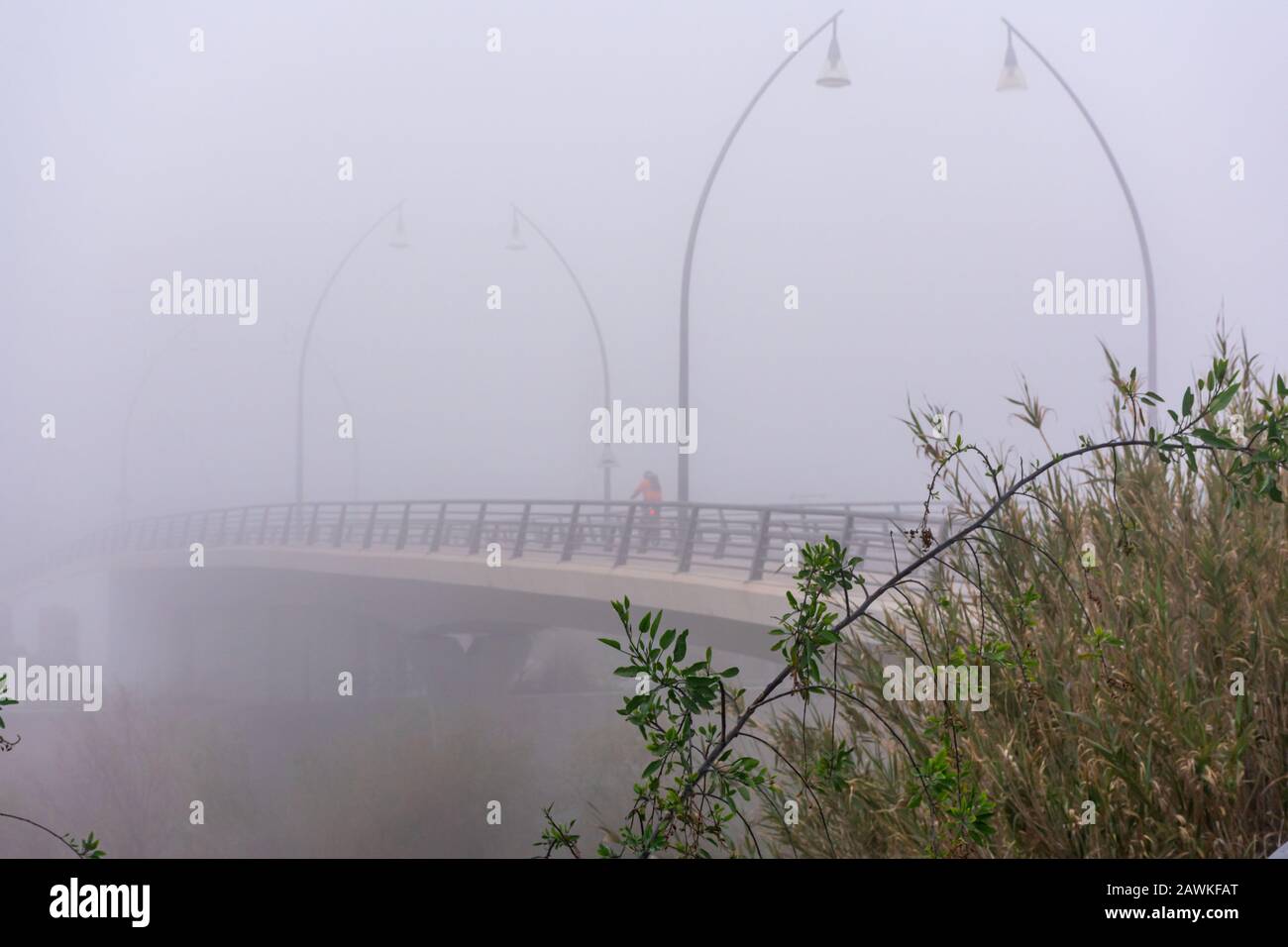 Giornata di nebbia su un ponte con lampade di strada che si perdono tra la nebbia e un ciclista che attraversa Foto Stock