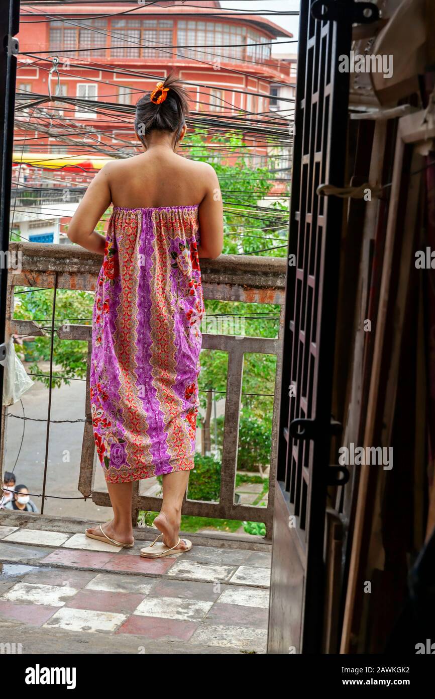 Una povera, povera donna cambogiana impoverita che indossa un colorato abito a sacco è in piedi su un balcone che guarda giù la strada a Kampong Cham, Cambogia. Foto Stock