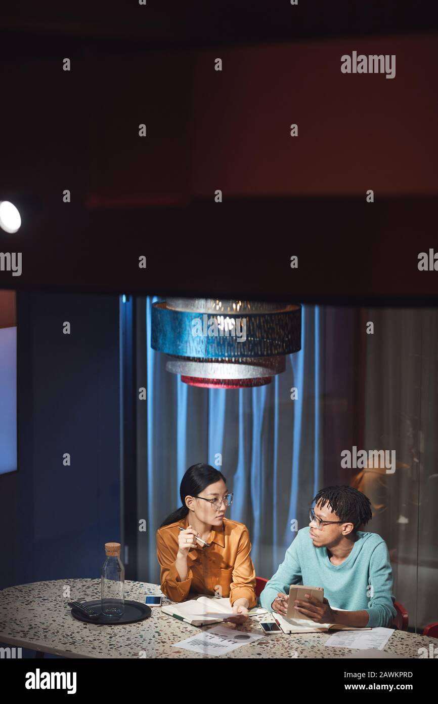 Ritratto ad alto angolo di due persone di affari etnici che discutono il progetto mentre lavora in ritardo in ufficio scuro, copia spazio Foto Stock