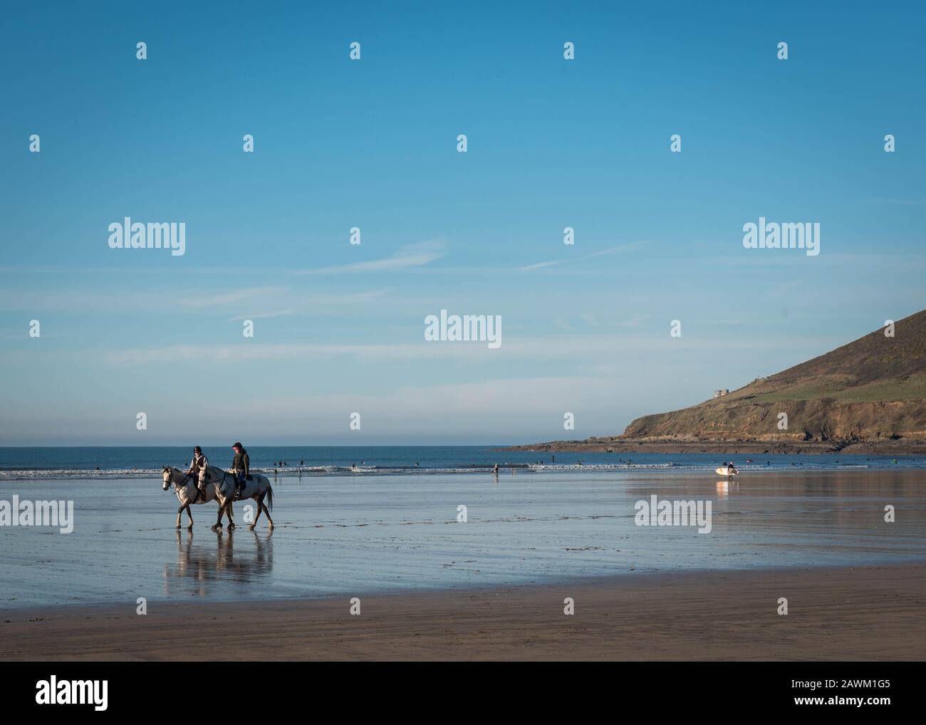 Coppia di cavalli che vengono cavalcati lungo la spiaggia di Saunton Sands nel Devon del Nord, Inghilterra al sole con i surfisti in background Foto Stock