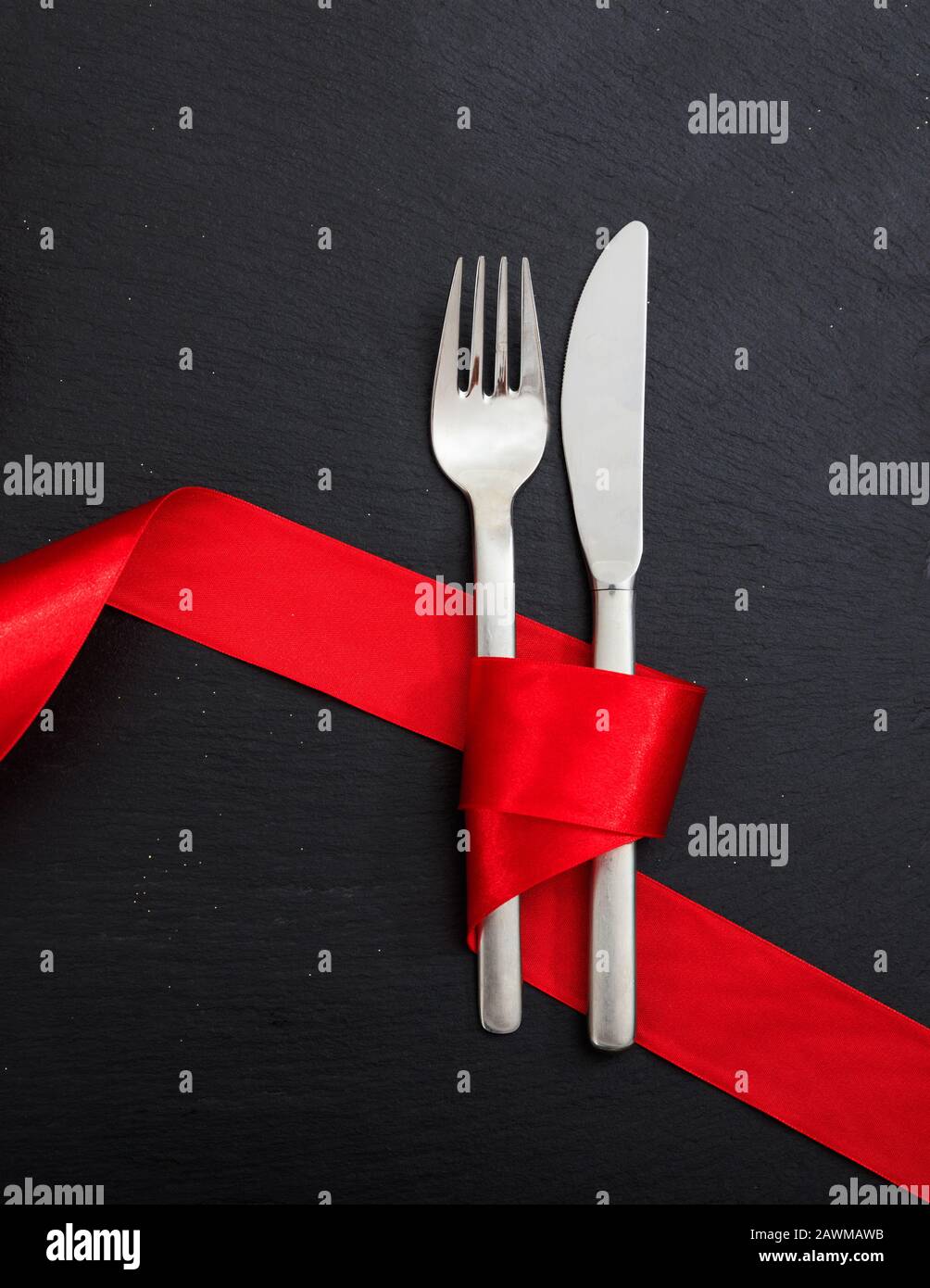 Cena di San Valentino con tavolo. Posate a forchetta e coltello legate con nastro rosso passione su sfondo nero, vista dall'alto. Foto Stock
