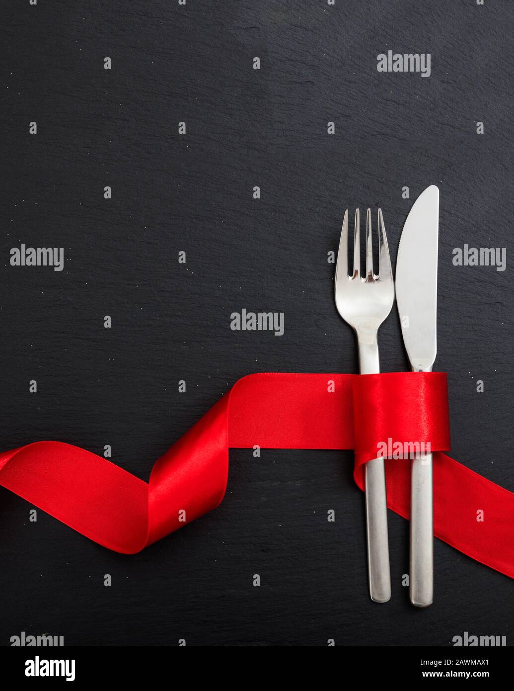 Cena di San Valentino con tavolo. Posate a forchetta e coltello legate con nastro rosso passione su sfondo nero, vista dall'alto. Foto Stock