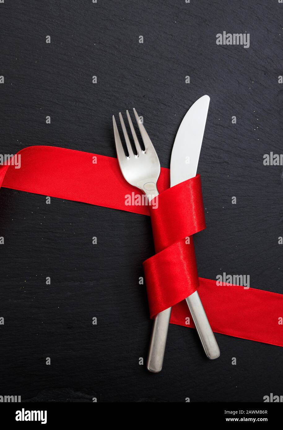 Cena di San Valentino con tavolo. Posate a forchetta e coltello legate con nastro rosso passione su sfondo nero, vista dall'alto. Foto Stock