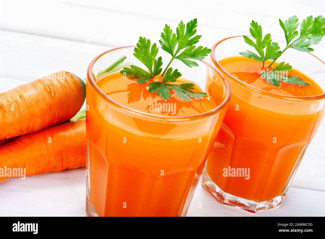 Concetto di cibo sano fatto in casa - due bicchieri di succo di carota fresco con prezzemolo accanto a tuberi di carote intere su un tavolo bianco di legno Foto Stock