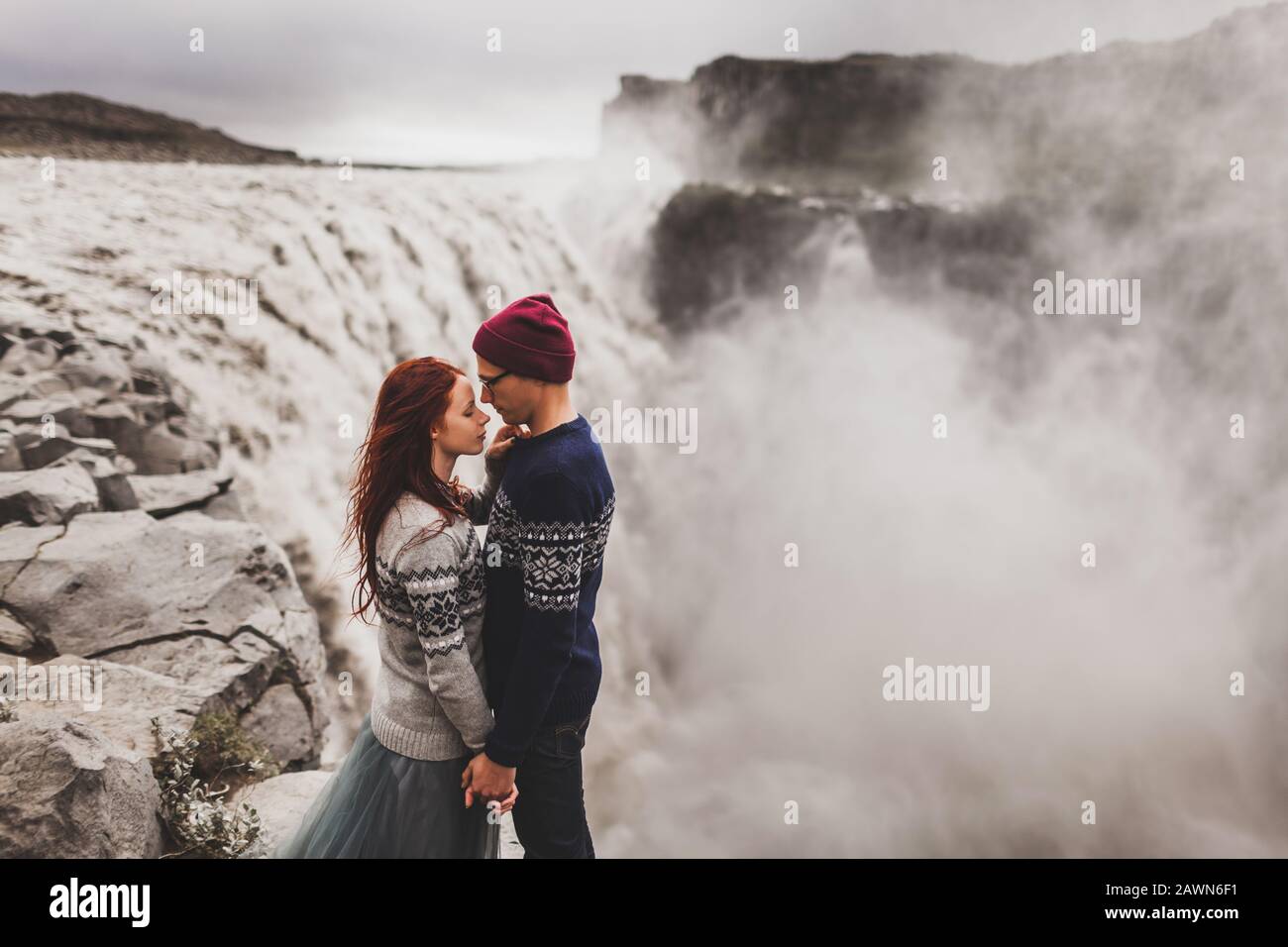 Giovane coppia elegante in amore kissing insieme vicino alla celebre paesaggio islandese di Dettifoss cascata. Tradizionale maglioni di lana, cappello, capelli rossi, mantello grigio. Drammatico paesaggio, freddo in Islanda. Foto Stock