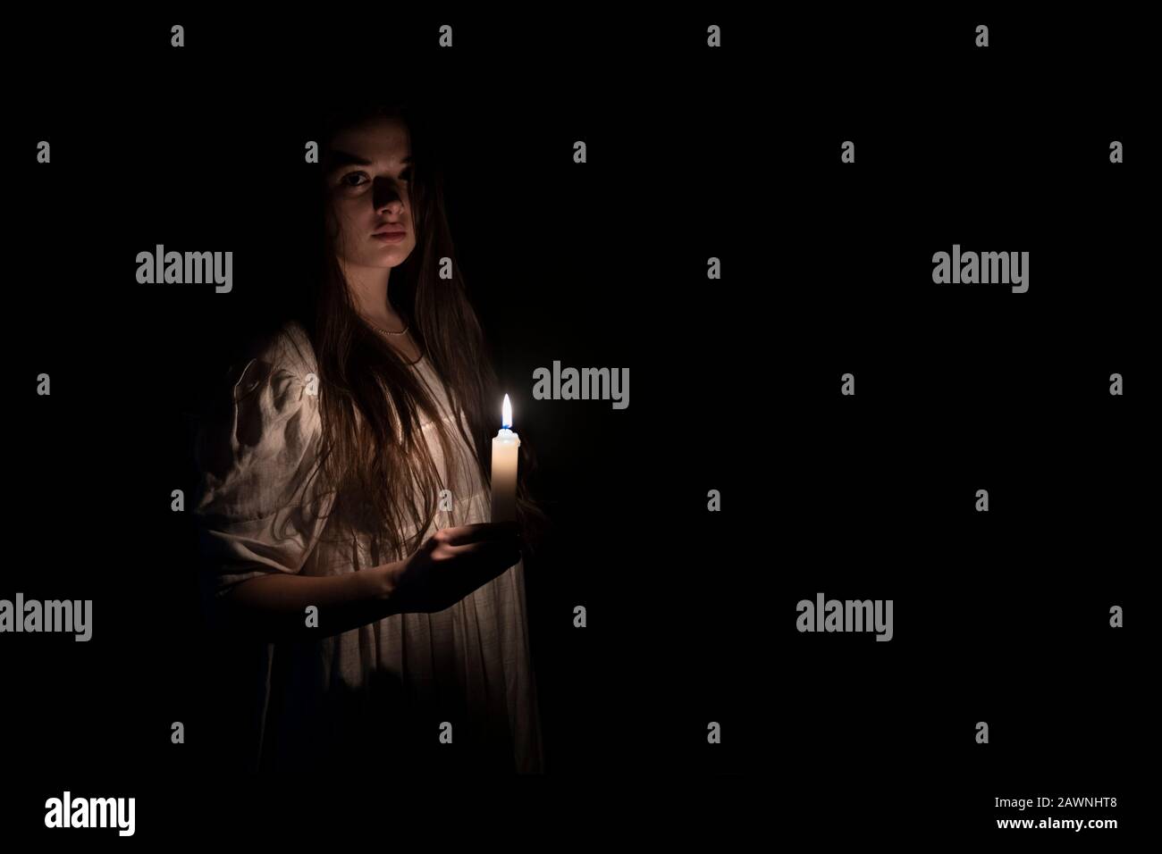 Una giovane ragazza in un vecchio abito bianco che tiene una candela in mano e fissando la macchina fotografica. Spazio di copia sul lato destro. Sfondo scuro. Foto Stock