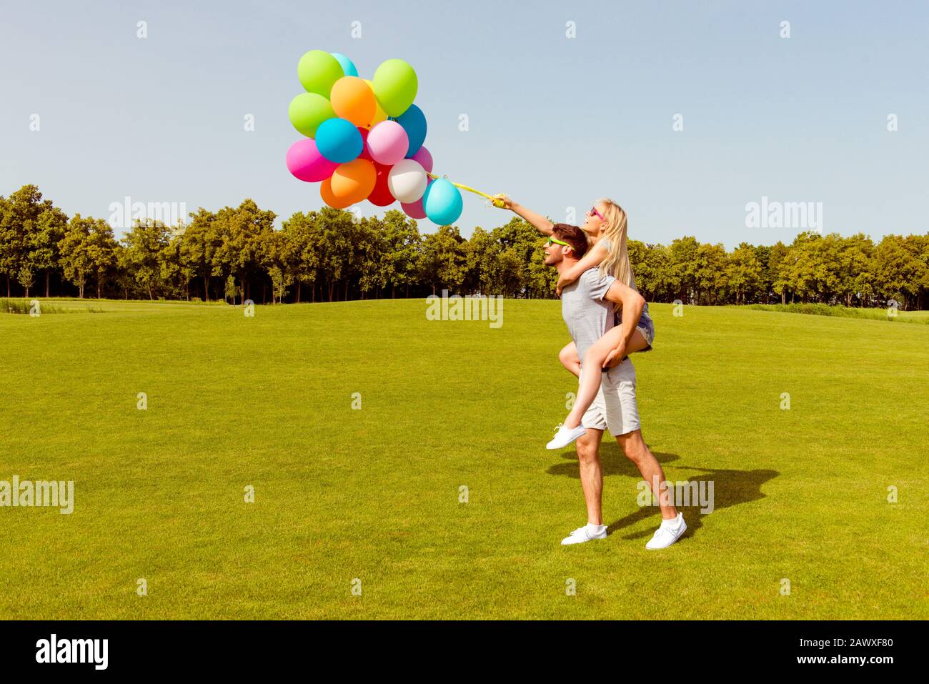 Giovane e simpatico uomo che cammina nel parco e che porta indietro la sua ragazza con palloncini Foto Stock