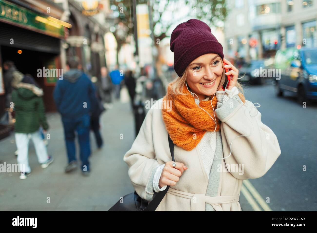 Giovane donna in cappellino e sciarpa che parla su smartphone sul marciapiede urbano Foto Stock