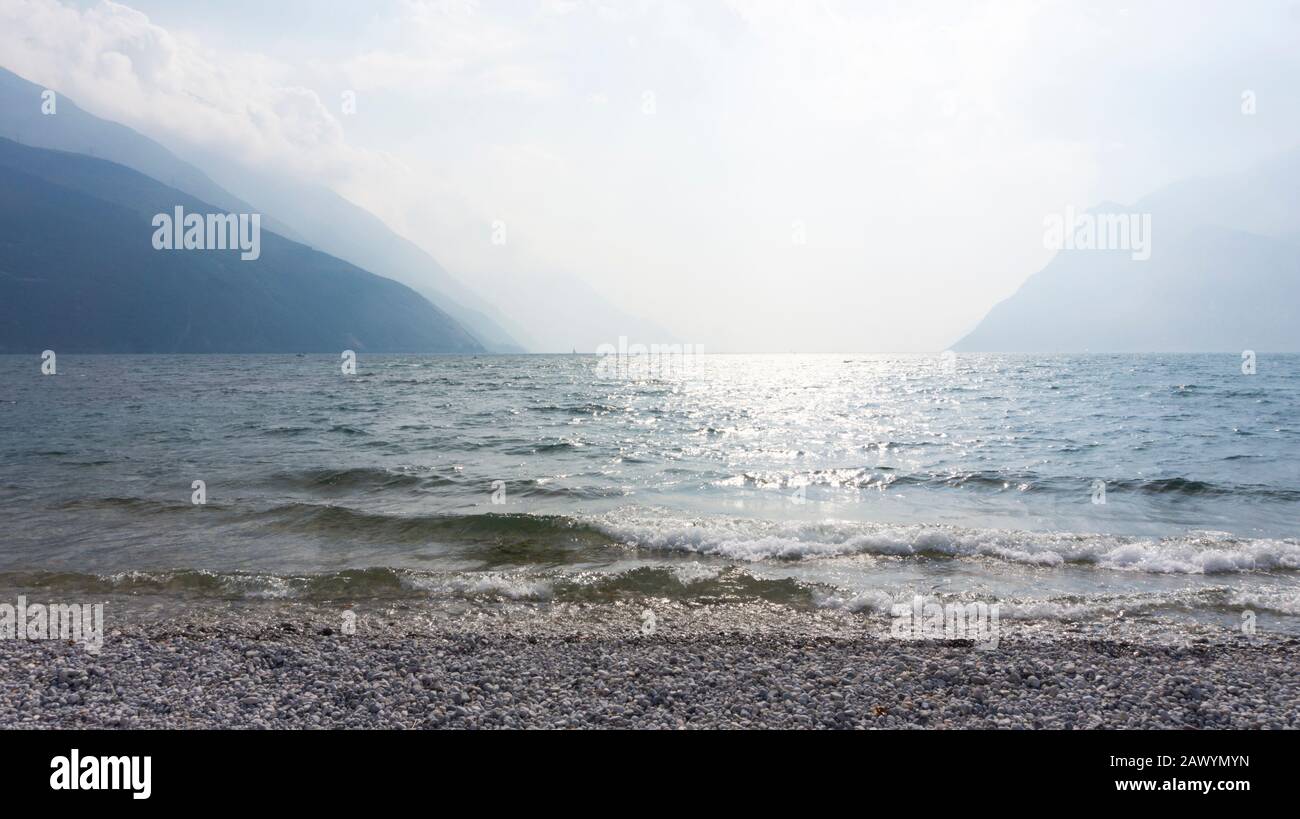 Vista panoramica sul lago di Garda da una spiaggia di ciottoli al sole e luce nebulosa. Rilassante paesaggio acquatico di onde luminose sulla superficie di un Foto Stock