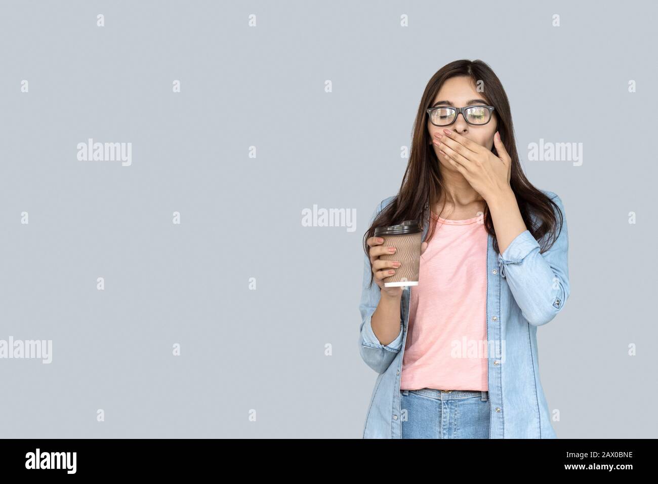 Giovane stanco sonno ragazza indiana sbadigia tenendo il caffè isolato sul grigio Foto Stock