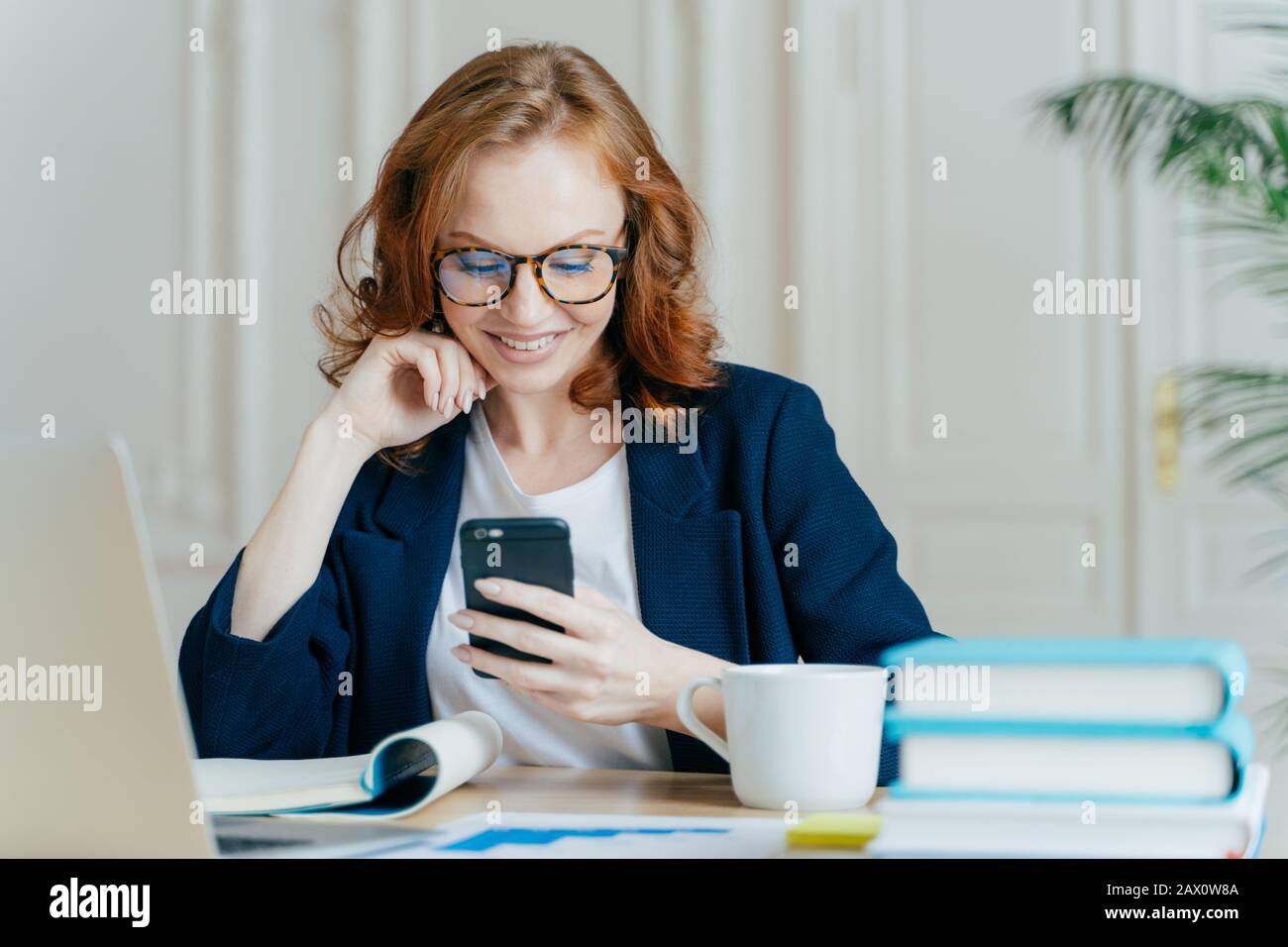 Foto di graziose donne si siede con dispositivo smartphone, tipi di feedback, lavora in ufficio su un laptop aggiornato, focalizzato sullo schermo del gadget, si siede sul lavoro Foto Stock