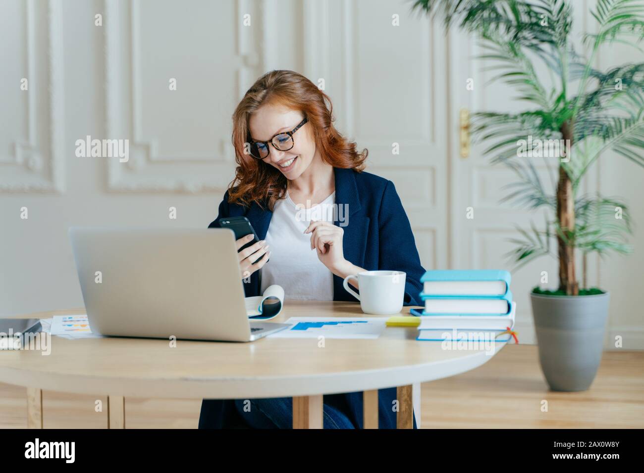 Sorridente femmina dai capelli rossi messo a fuoco nel cellulare moderno, felice di ricevere il messaggio di testo, pose sul posto di lavoro, bevande caffè, lavori sul progetto finanziario GET Foto Stock