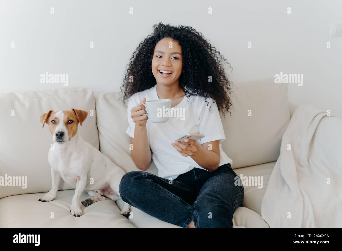 La donna con pelle scura e deliziata dispone in un appartamento moderno, si siede su comodo divano con animale domestico, bevande caffè, usa il telefono cellulare per la comunicazione online, Foto Stock