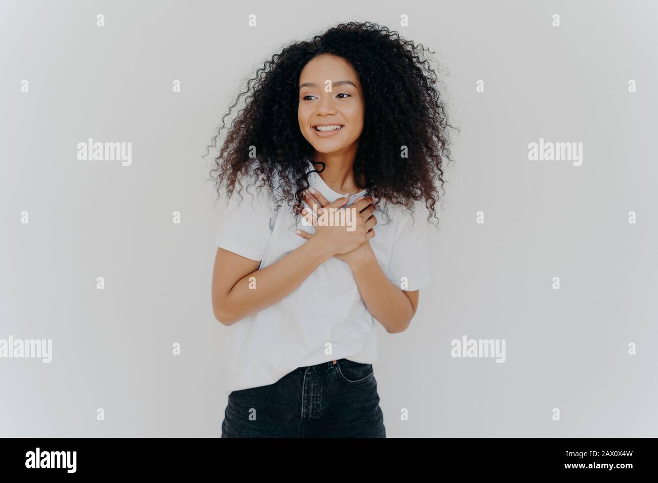 La foto della ragazza felice tiene entrambe le mani sul petto, guarda da parte con sorriso piacevole, nota qualcosa di impressionante, pone contro sfondo bianco, indossa casua Foto Stock