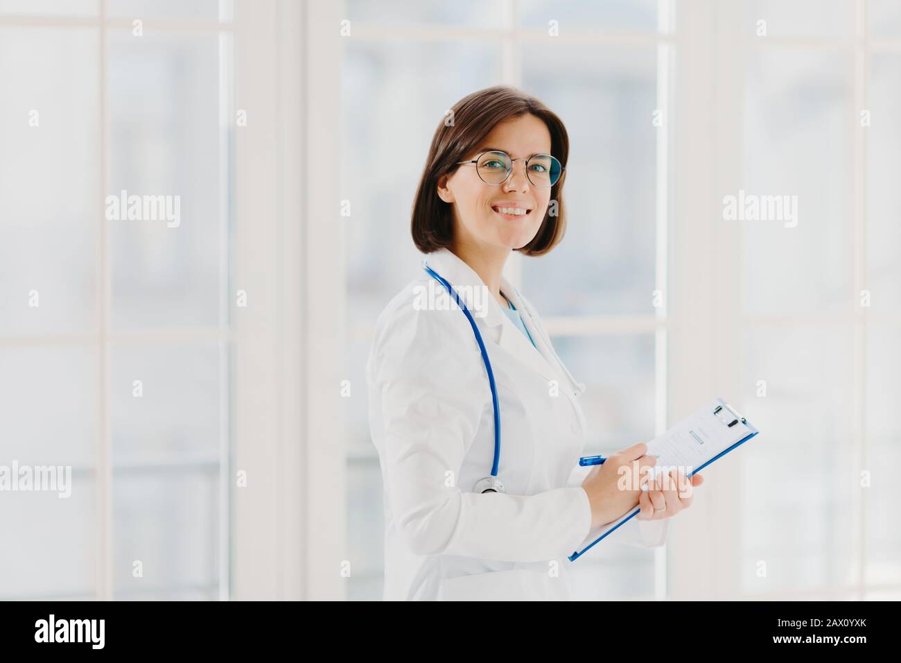 Il colpo orizzontale del medico femminile riempie in su la forma medica alla clipboard, si alza all'interno, indossa gli occhiali rotondi, il gown bianco e lo stetoscopio. Pratica generale Foto Stock