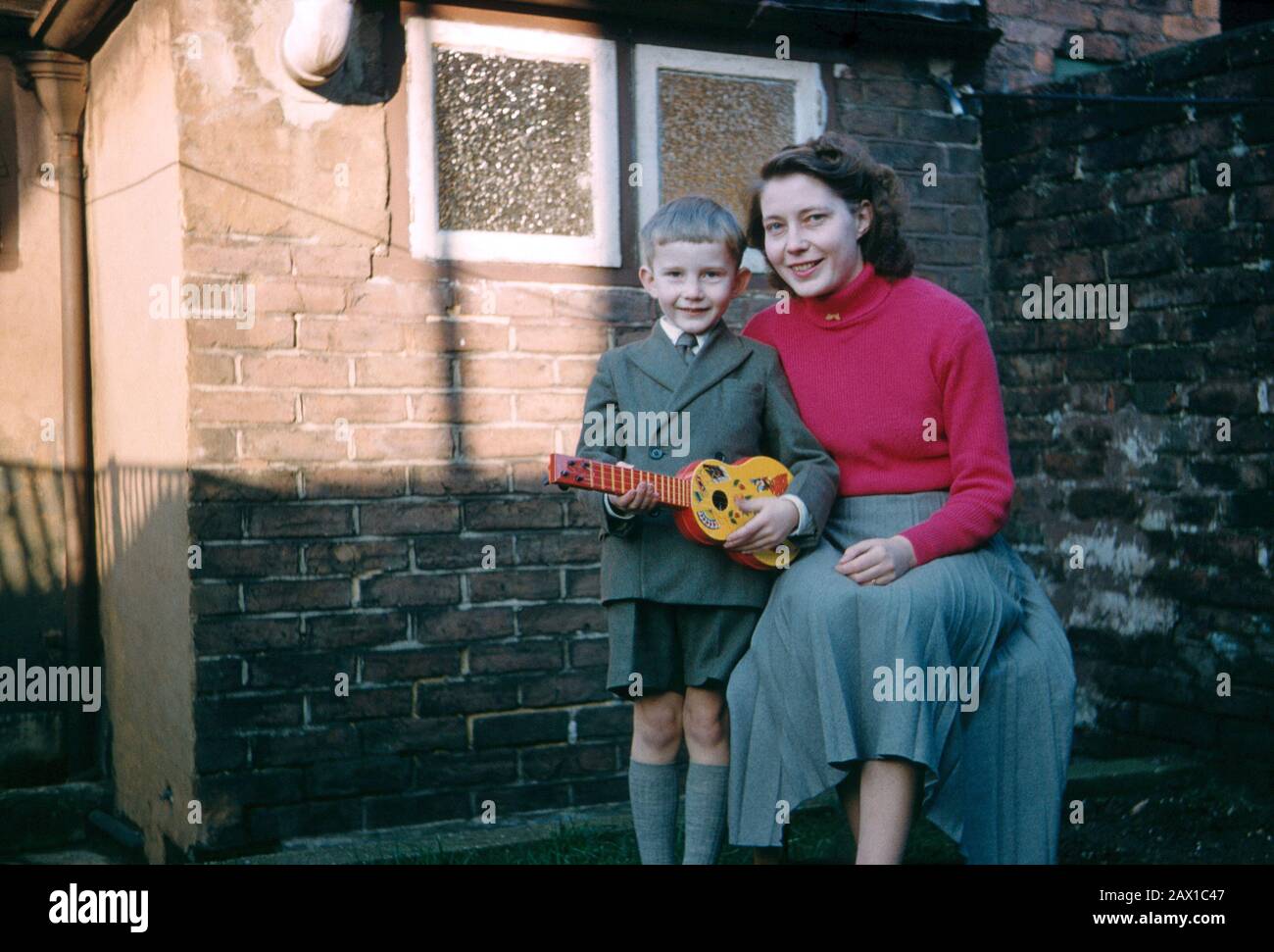 Ragazzo di cinque anni che indossa una tuta corta a due alberi e tiene una chitarra giocattolo che posa con sua madre nel cortile posteriore di una casa a schiera Foto Stock