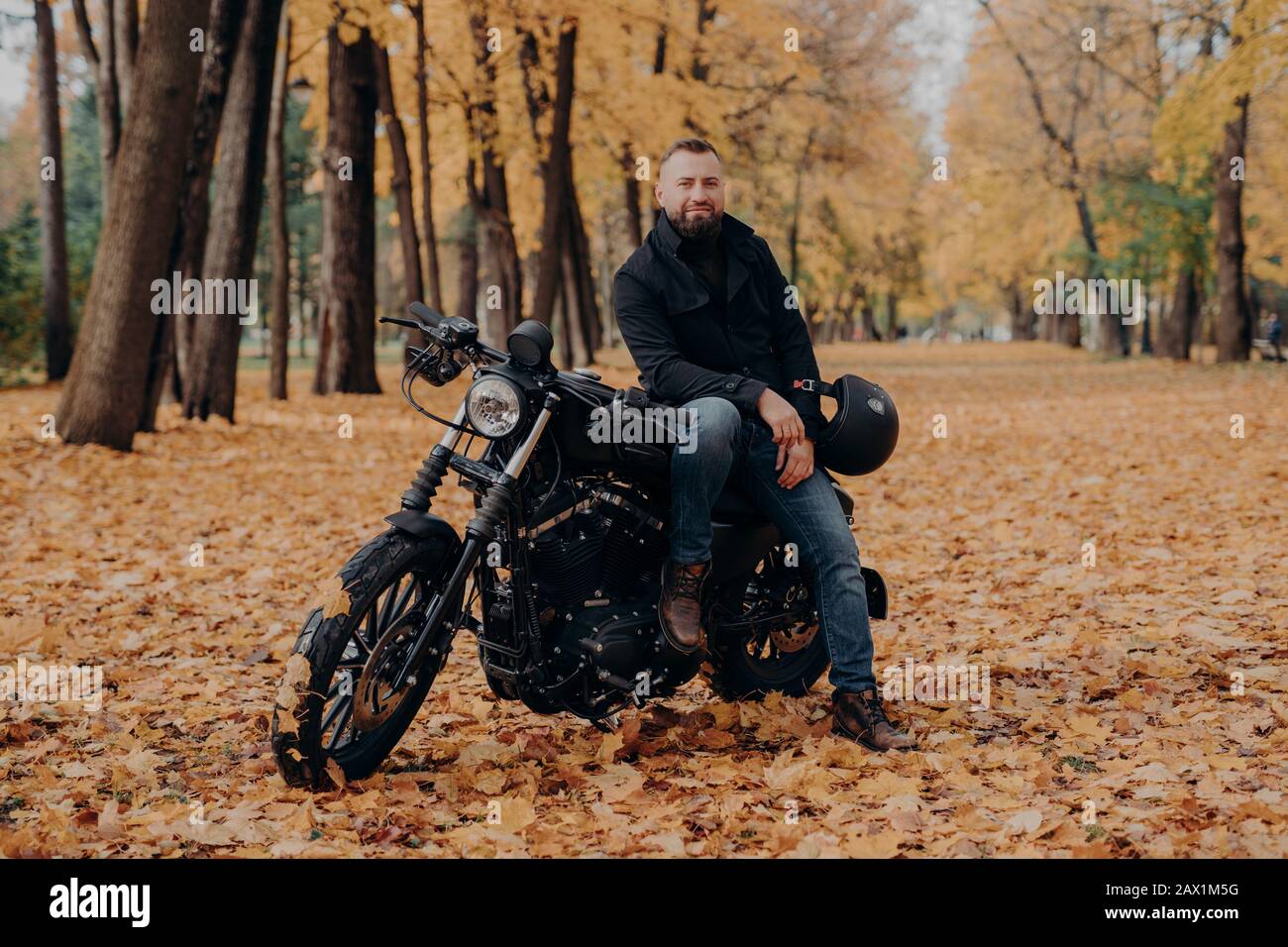 Il biker con bearded dispone su propria motocicletta, tiene il casco, guida la moto, si dispone all'aperto nel parco, terra coperta con foglie cadute. Stile di vita biker. Adven Foto Stock