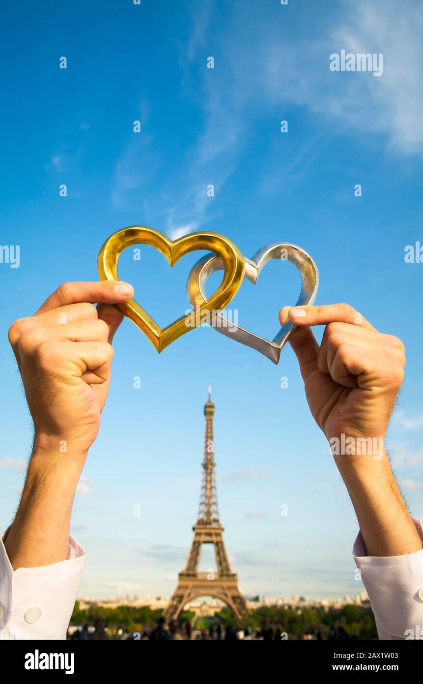 Mani che tengono un paio di cuori d'oro e d'argento all'aperto davanti a una vista soleggiata della Torre Eiffel a Parigi, Francia Foto Stock