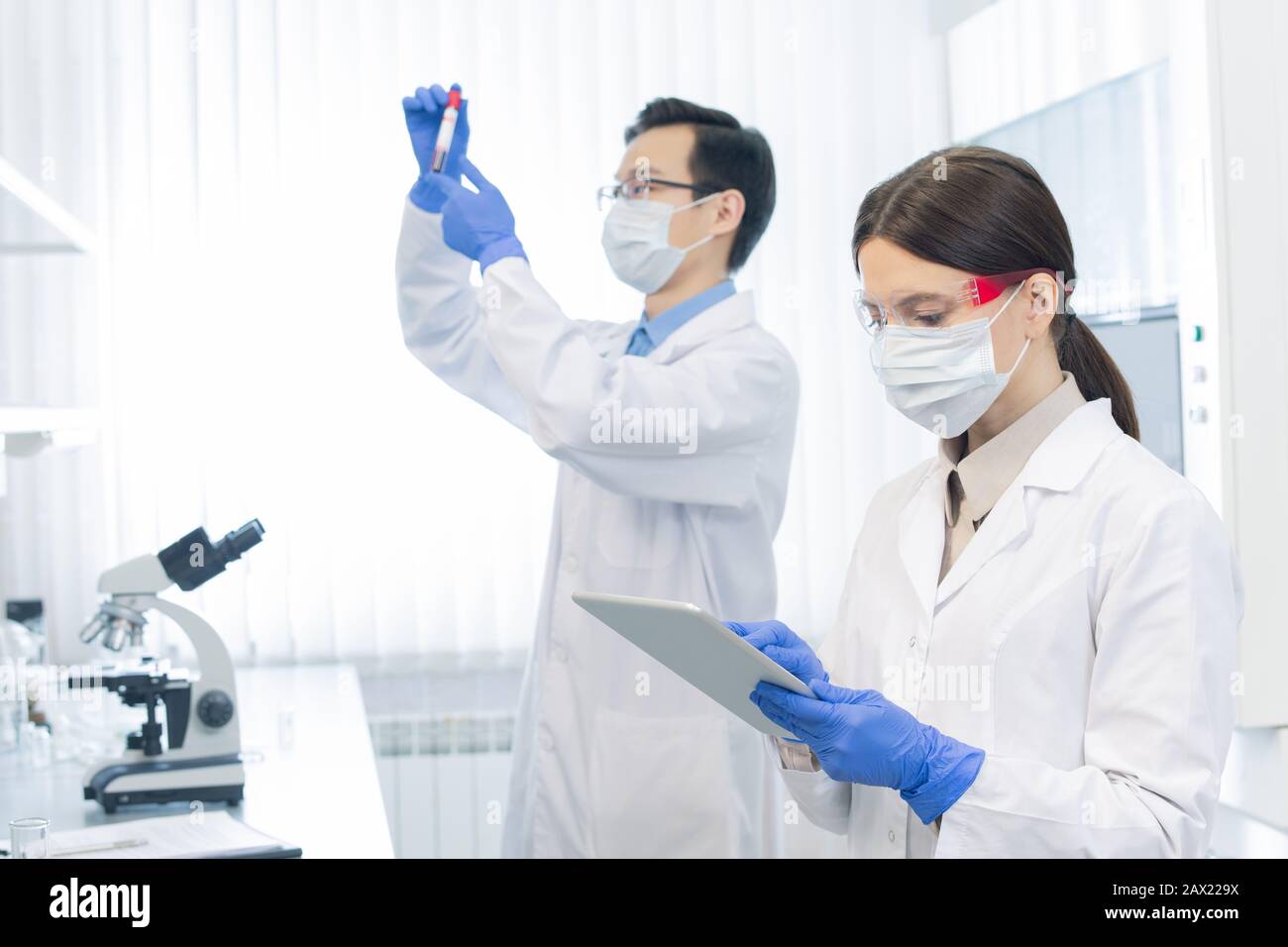 Due giovani scienziati che indossano maschere protettive e guanti che lavorano su nuovi farmaci in moderno laboratorio Foto Stock